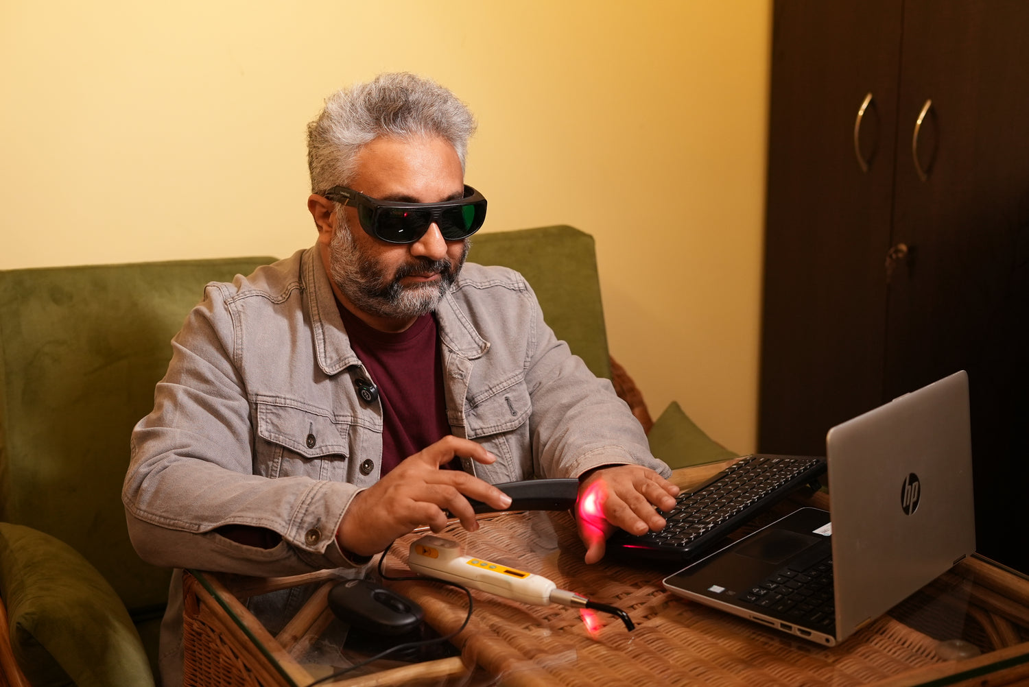 Man wearing sunglasses and a gray jacket using a laptop on a couch.